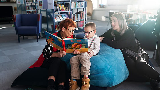 Volontären och läsfaddern Lena Olsson och Eman Begic sitter på kuddar och läser en bok. Bredvid sitter Emans mamma Alma.