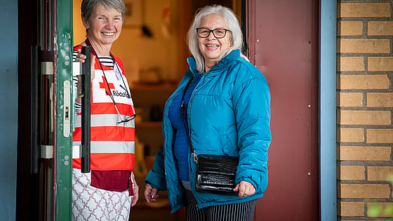 En kvinnlig volontär i Röda Kors-väst håller upp dörren för en kvinnlig stödsökande i blå täckjacka.